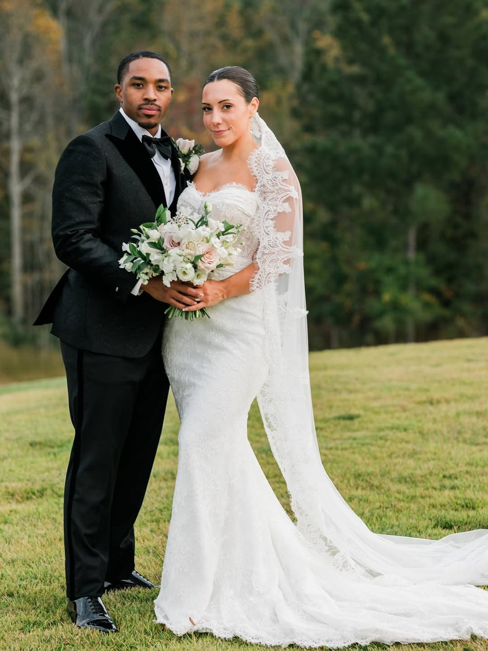 A bride in a fitted white lace wedding gown with a long sheer lace veil and a bouquet of white and blush flowers, standing outdoors with a groom in a black Dionicios Closet custom tuxedo on a grassy lawn with trees in the background.