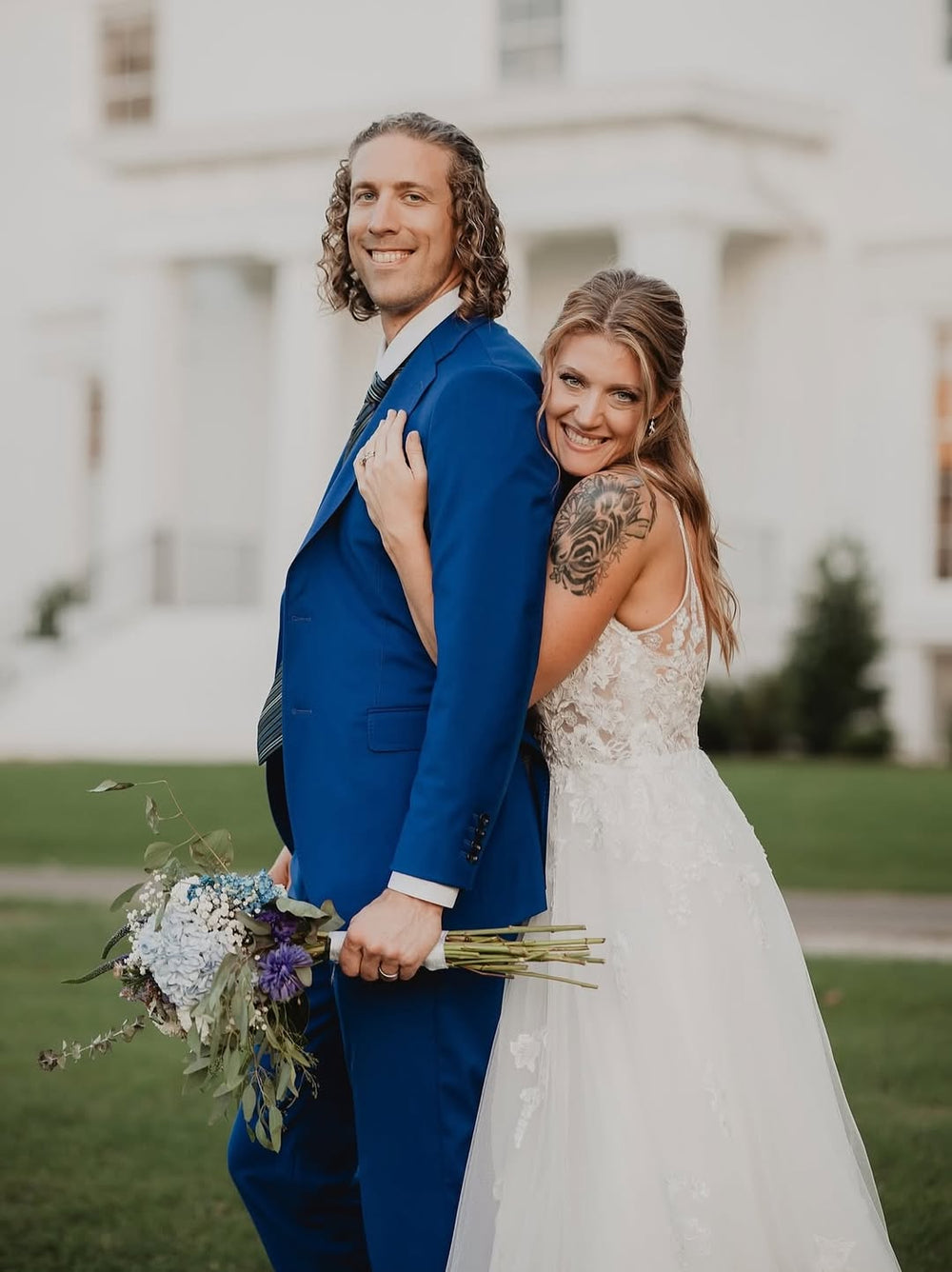 A smiling couple outdoors in front of a white building. The man wears a bright blue Dionicios Closet suit and holds a bouquet, while the woman wears a white lace wedding dress and leans against him.