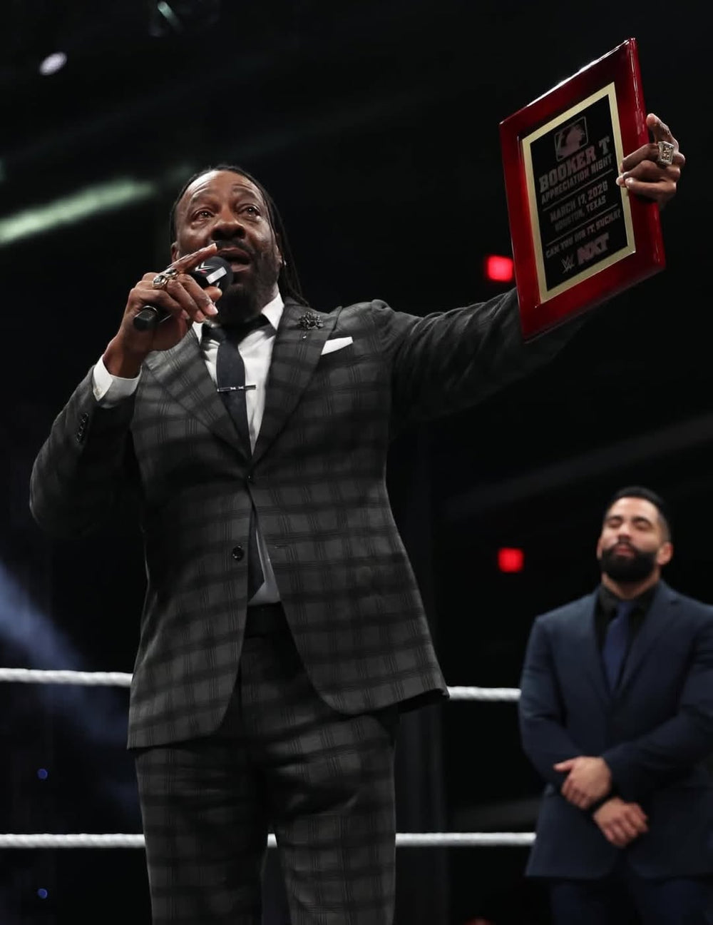 A man in a custom grey and black plaid Dionicios Closet suit, stands inside a boxing ring holding a microphone and a framed award plaque