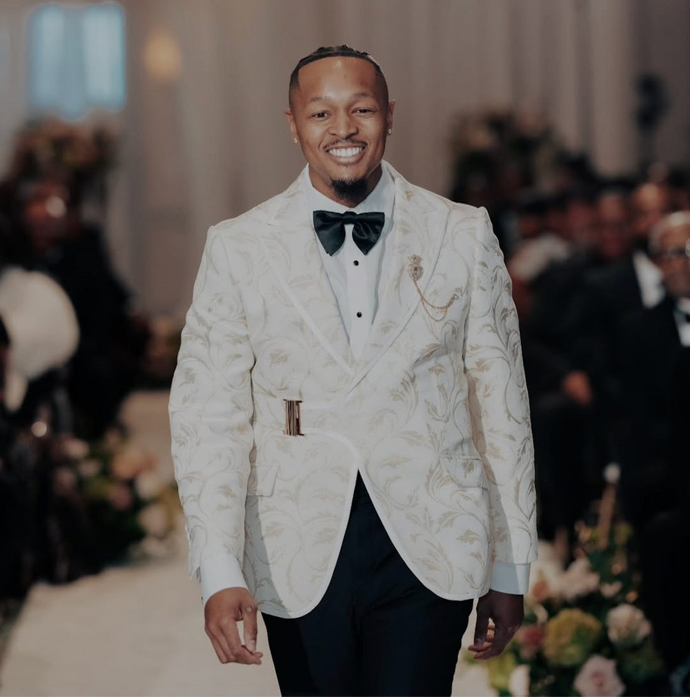 A smiling man walks down a runway wearing a white Dionicios Closet custom tuxedo jacket with ornate gold floral embroidery, a white shirt, a black bow tie, and black trousers. The background is a softly blurred indoor event setting with seated guests.