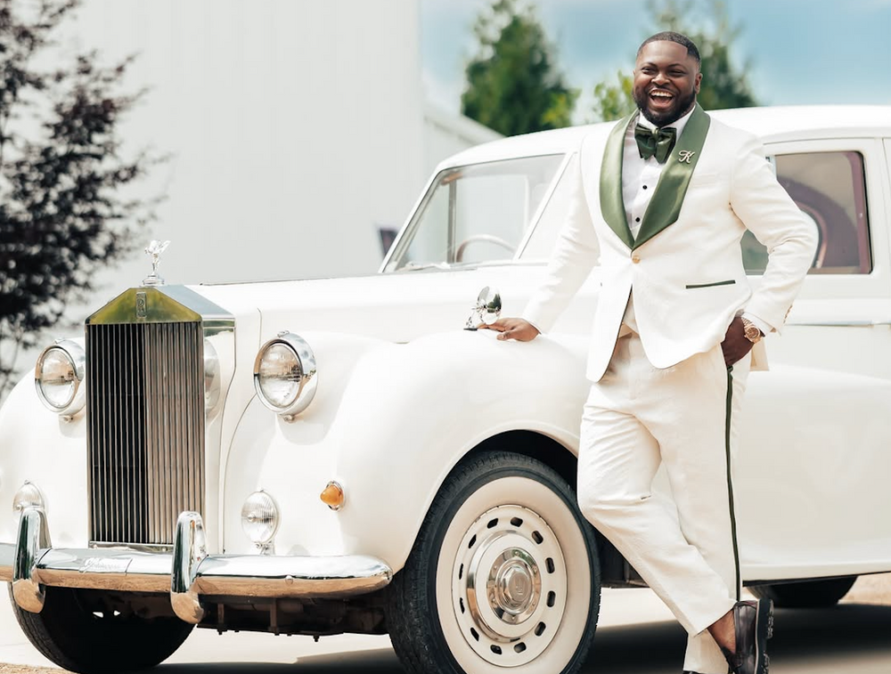 A man in a Dionicios Closet custom white tuxedo with green accents leans against a vintage white Rolls-Royce car outdoors.