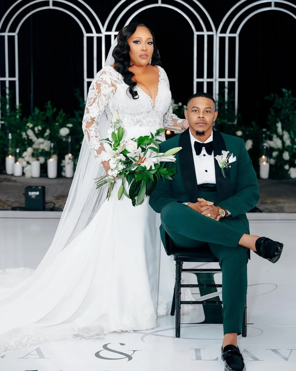 A bride in a white lace wedding gown holding a bouquet of white flowers stands next to a man in a dark green custom Dionicios Closet tuxedo with a black bow tie and boutonniere, posed in front of a black arched backdrop with white candles and greenery.