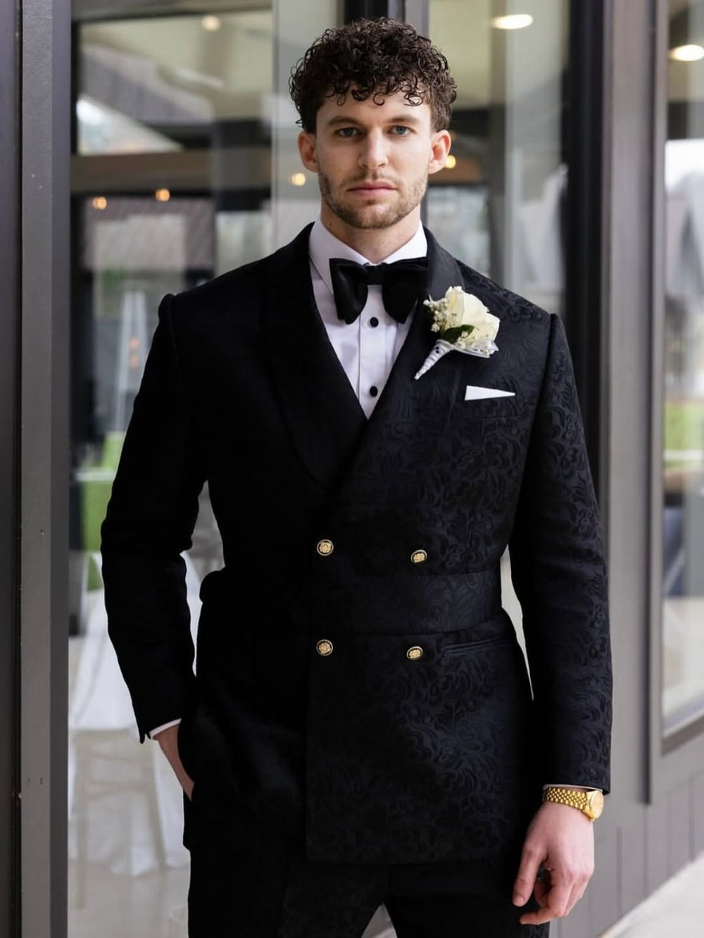 A man wearing a Dionicios Closet custom black double-breasted tuxedo with a subtle damask pattern, a black bow tie, a white dress shirt, a white boutonniere, and a white pocket square, standing in front of a glass door.