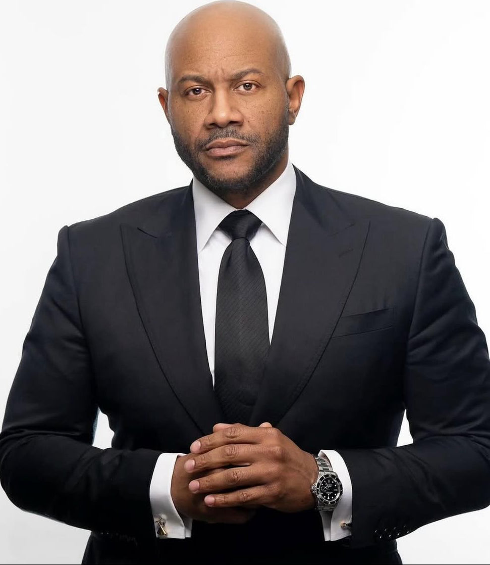 A professional portrait of a bald man with a beard, wearing a black suit, white dress shirt, and black tie, standing against a solid white background.