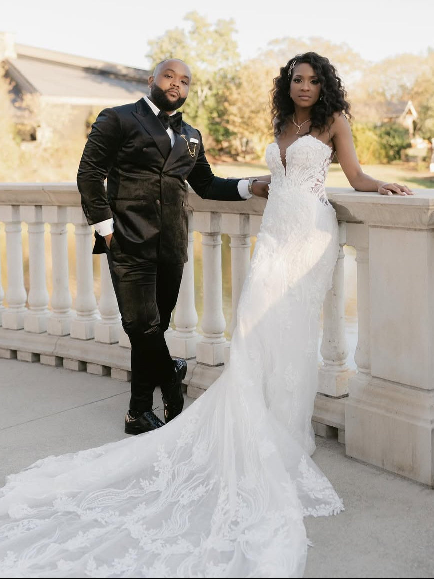 A bride in a strapless white lace wedding gown with a long train stands on a stone terrace beside a groom in a black Dionicios Closet tuxedo, with a sunny outdoor background of trees and houses.