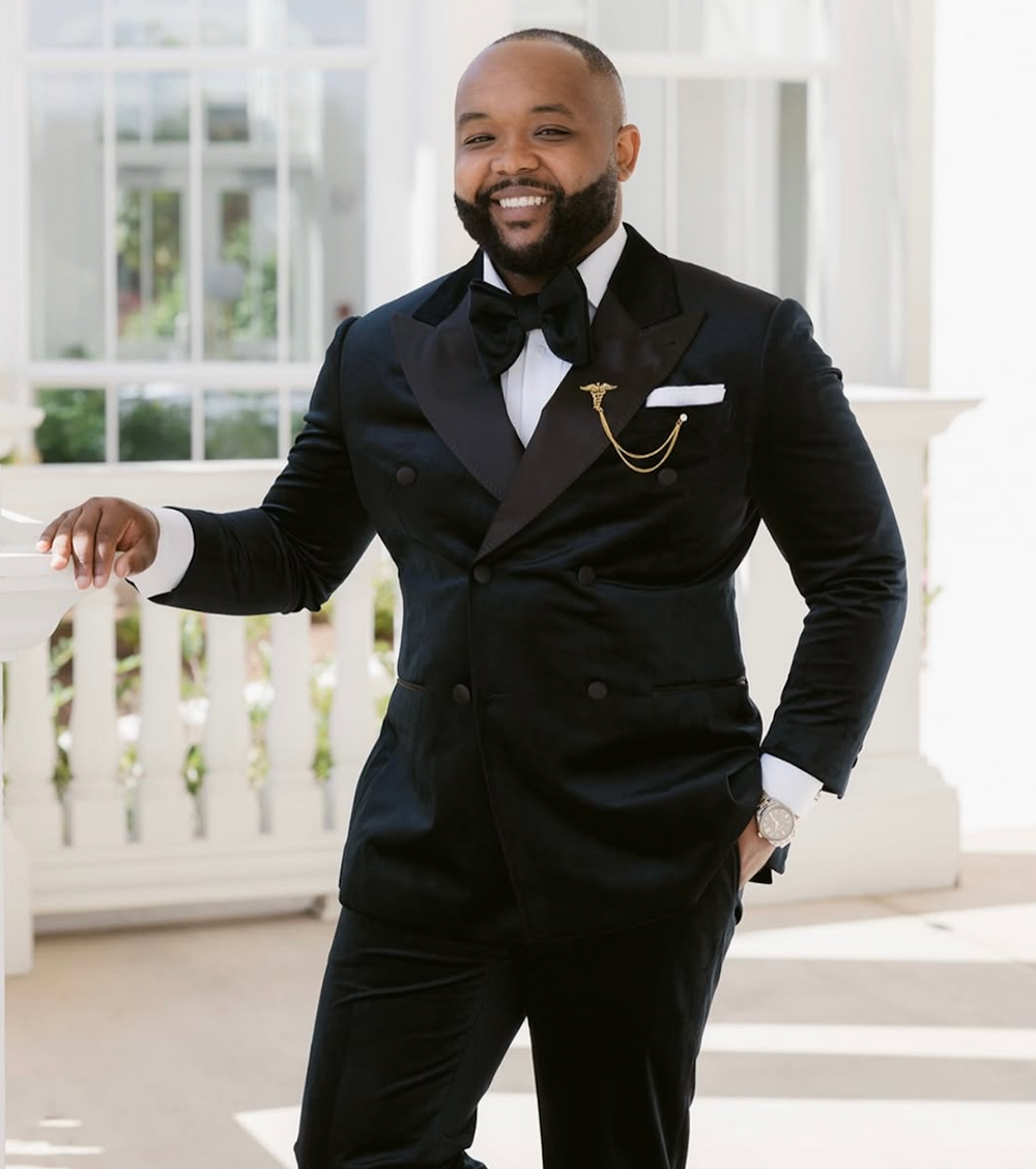 A smiling man wearing a black double-breasted Dionicios Closet custom tuxedo with a black bow tie, white dress shirt, and gold pocket square, standing on a white porch.