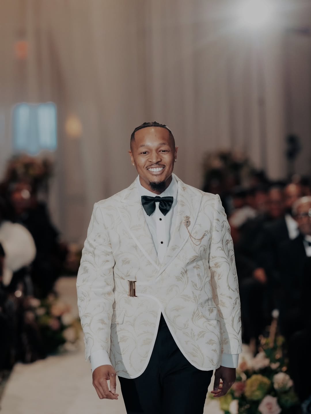 A man walking down a runway wearing a white patterned custom dionicios closet tuxedo jacket with gold detailing, a black bow tie, and black trousers, with a blurred indoor event background.