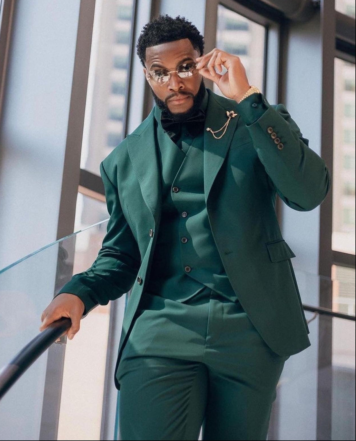 A man wearing a dark green Dionicios Closet three-piece custom suit with a matching vest and trousers, styled with a black shirt and gold jewelry, posing on an indoor staircase with large windows in the background.